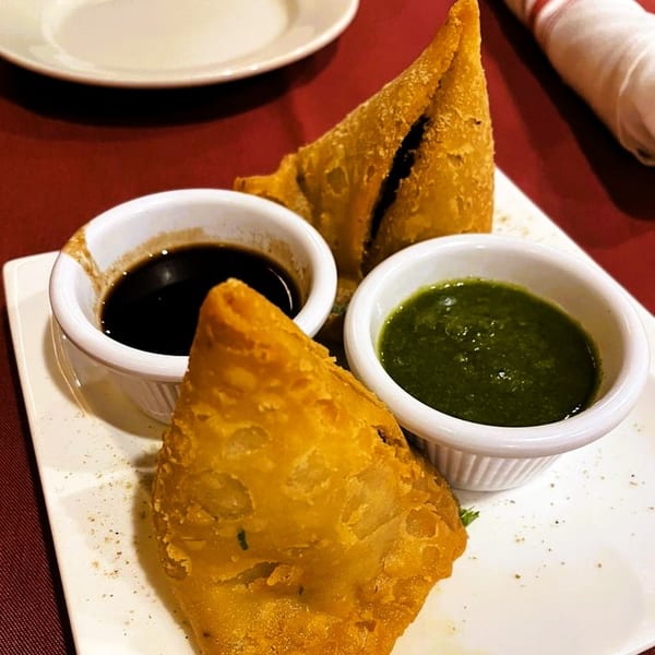 Two golden-brown samosas with dark and green dipping sauces on a white plate at Taj Indian Restaurant.
